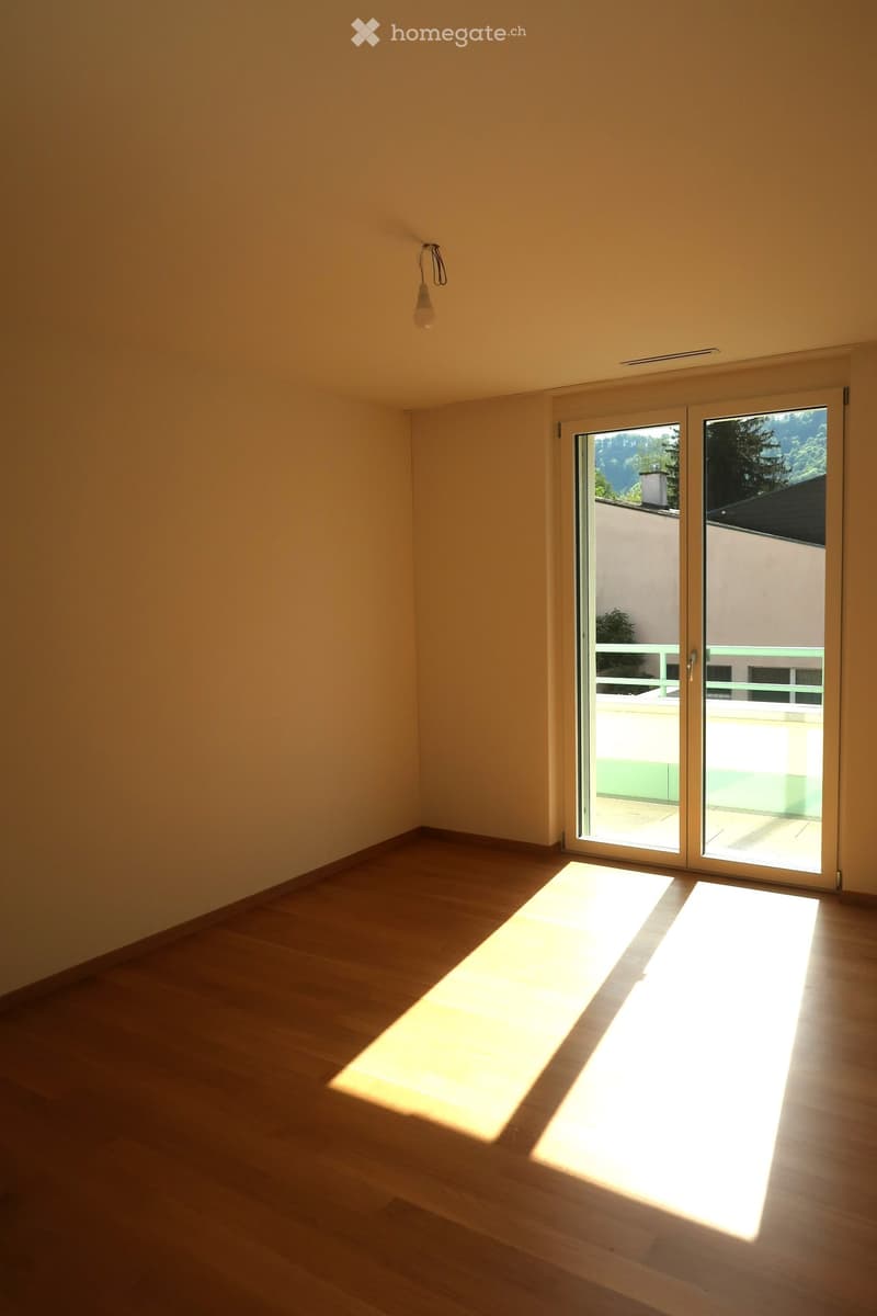Empty room with wooden flooring and large sliding glass doors leading to a balcony. Sunlight streams in, casting shadows on the floor.