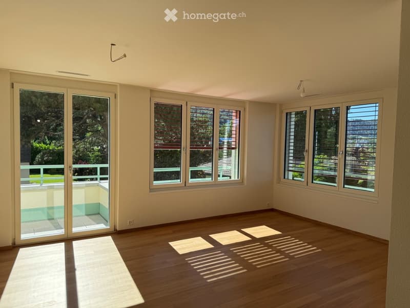 Empty room with wooden flooring, large windows, and sliding glass doors leading to a balcony. Natural light fills the space.