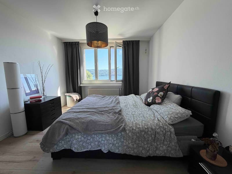 Modern bedroom with a large bed, nightstand, mirror, and a view of the city through large windows.