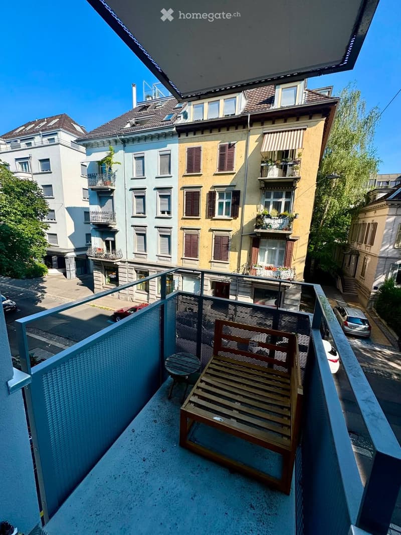 Balcony with a wooden bench and a small table, overlooking a street with multi-story residential buildings and parked cars.