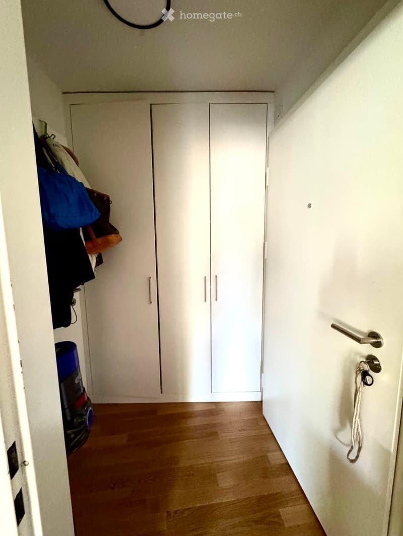 A narrow hallway with wooden flooring, white walls, and built-in white closet doors. A vacuum cleaner and some clothing are visible on the left side.