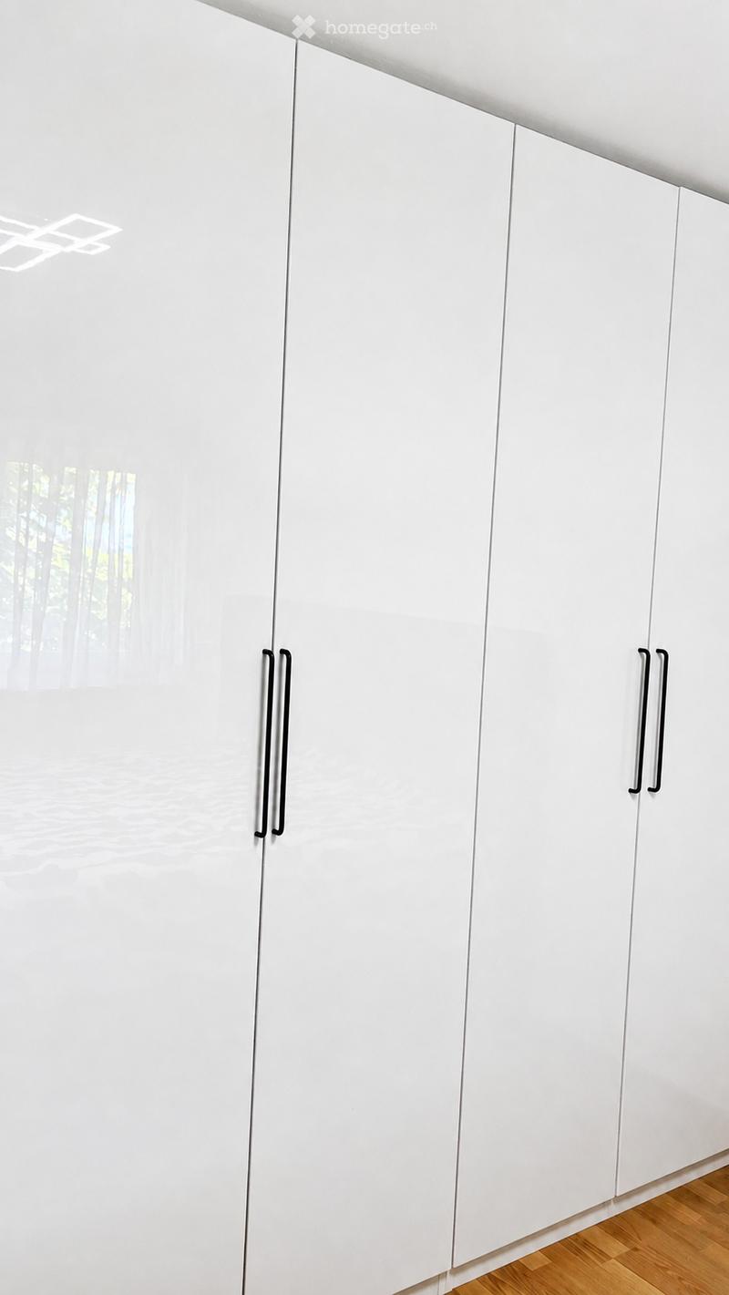 Modern kitchen with white cabinetry and sleek black handles, featuring a wooden floor and a view of greenery through a window.