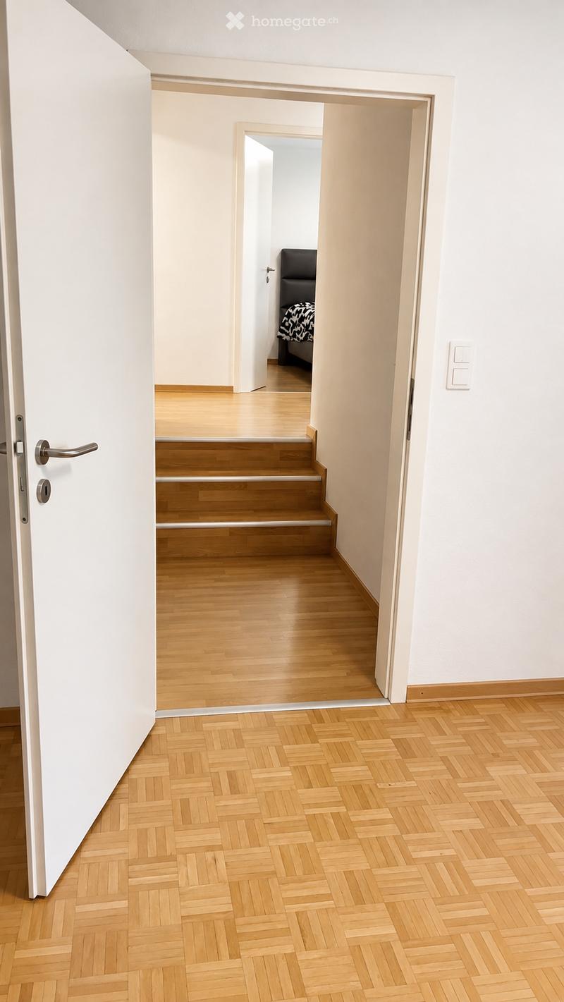 A clean, modern hallway with light wooden flooring and white walls. The hallway features a staircase leading upwards, with a door on the left and a glimpse of a bedroom visible at the end of the corridor.