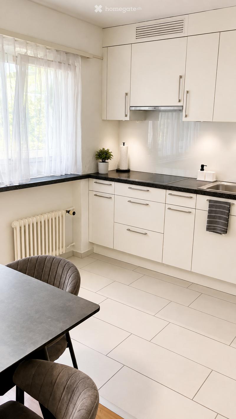 Modern kitchen with white cabinets, black countertops, and stainless steel appliances. Features a dining area with a grey table and chairs, a sink, and a small potted plant on the windowsill.