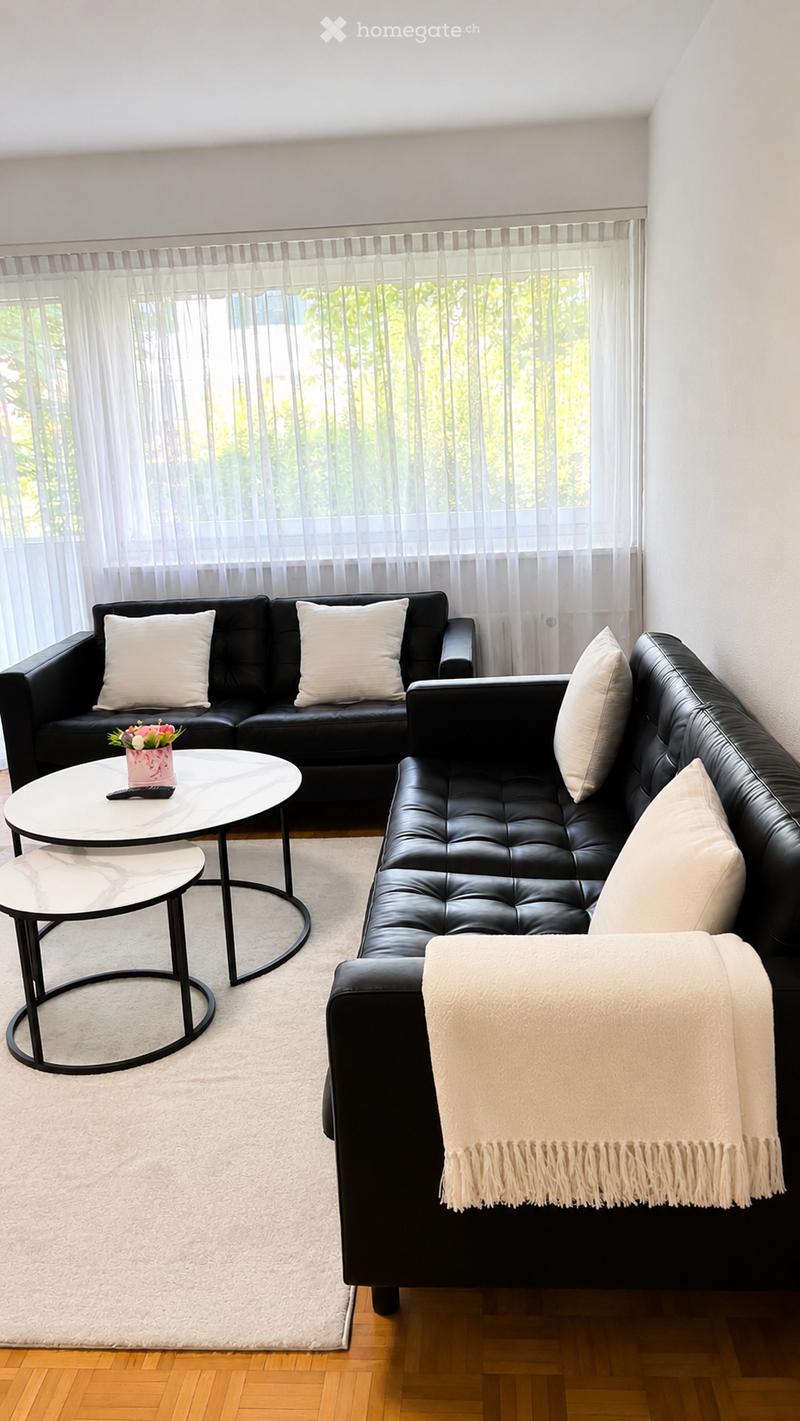 Modern living room with a black leather sectional sofa, white throw pillows, a white coffee table, a small round stool, a throw blanket draped over the sofa, and a small flower vase on the coffee table. The room features large windows with sheer curtains
