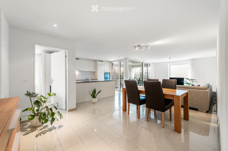 Bright, open-plan living and dining area with modern furniture, large windows, and a connected kitchen.