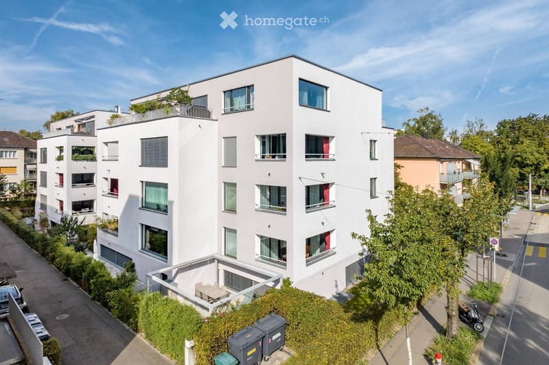 Modern white apartment building with multiple floors, balconies, and large windows. The building is surrounded by greenery and trees, with a small outdoor seating area visible on the ground floor.