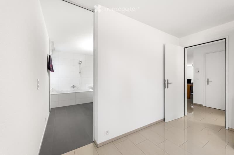 Modern bathroom with white tiled walls, a bathtub, a shower area, and a towel hanging on the wall. The floor is tiled in dark grey and light beige.