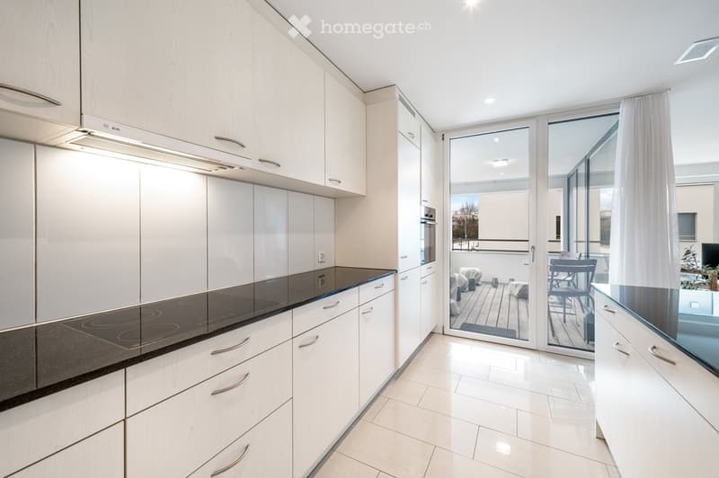 Modern kitchen with white cabinets, induction cooktop, and an oven. Large sliding glass doors open to a balcony with outdoor furniture.