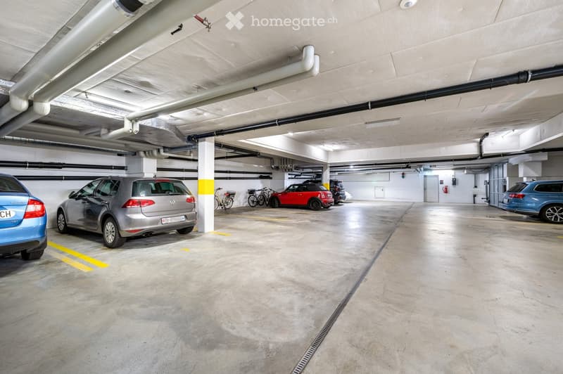 Underground parking garage with several parked cars, bicycles, and structural columns. The space is clean and well-lit with concrete floors and walls.