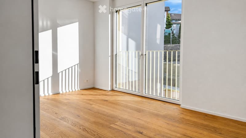 Empty room with wooden flooring, white walls, large glass sliding doors leading to a balcony, and a white railing visible.