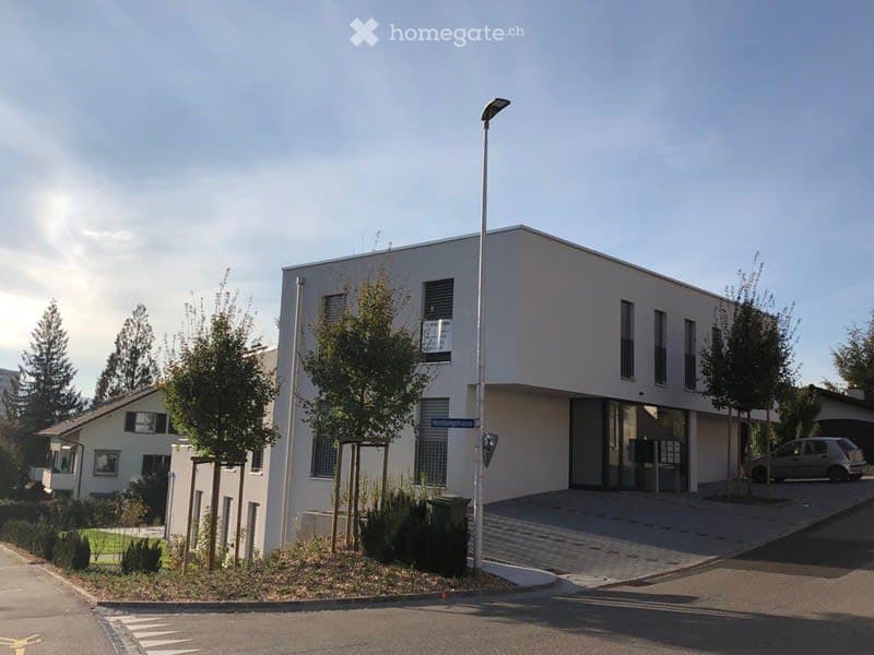 Modern, single-story building with a flat roof and white facade. The structure features large windows and a glass entrance. There are small trees and shrubs around the building, and a street lamp is visible in the foreground.