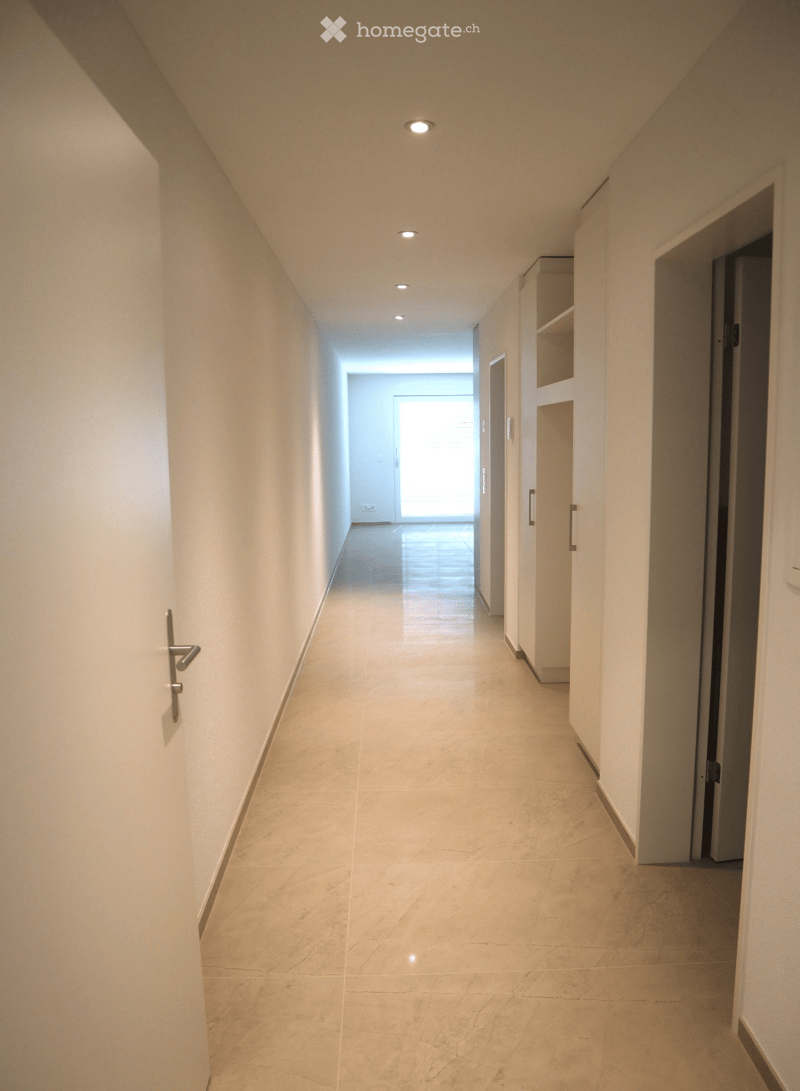 A clean, modern hallway with light-colored marble tiles, white walls, and recessed ceiling lights. The corridor features built-in shelving units and doors leading to other rooms.