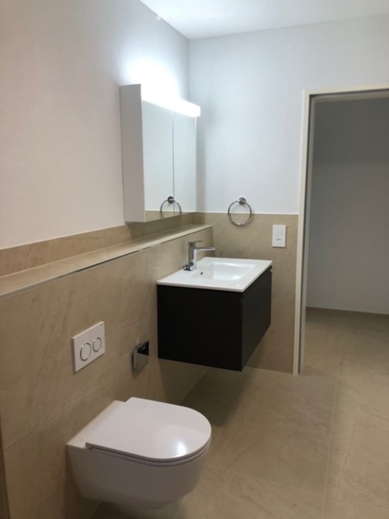 Modern bathroom with a white sink, sleek faucet, large mirror, and a toilet. The walls are tiled in beige, and the floor is covered with light brown tiles.