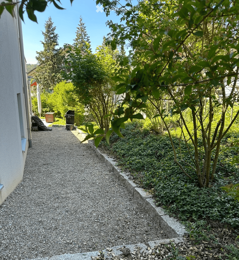 A narrow gravel pathway bordered by low stone walls, leading to a lush garden area with trees and shrubs. The pathway is flanked by greenery and small plants, with a covered outdoor area visible in the background.