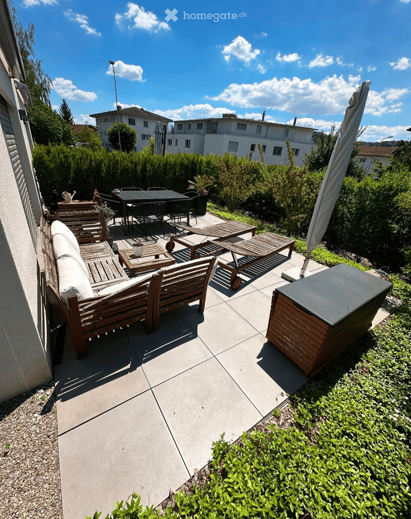 Balcony with wooden furniture including a sofa, table, chairs, and sun loungers, surrounded by greenery and a clear sky.