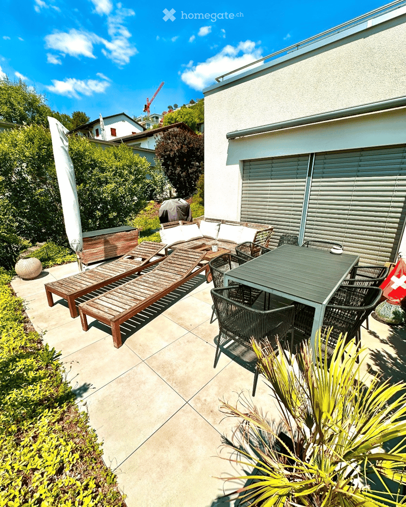 A well-maintained outdoor patio area with a sofa, lounge chairs, a dining table, and chairs, surrounded by greenery and a modern building.