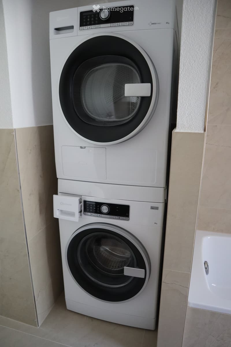 Modern white washing machine with digital control panel, placed in a tiled bathroom area.