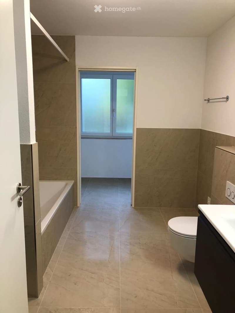 Modern bathroom with beige tiles, a bathtub, toilet, sink, and a window with frosted glass. The space is clean and well-lit.