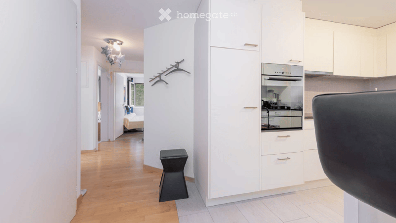 Modern, minimalist kitchen area with white cabinets, a built-in oven, a black stool, and a hanging coat rack. The floor is light wood, and the space is bright with natural light from a nearby window.