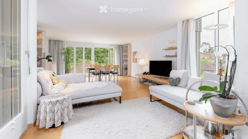 Bright, modern living room with white sofas, coffee tables, a TV, and large windows overlooking greenery.