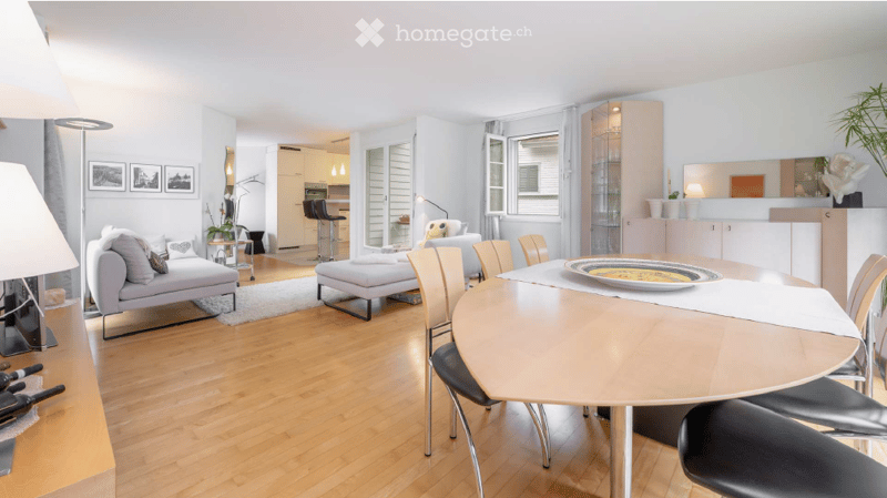 Bright, open-plan living and dining area with light wood flooring, white walls, and large windows. Features a grey sofa with cushions, an ottoman, a round dining table with chairs, a floor lamp, and a small kitchen area with bar stools.