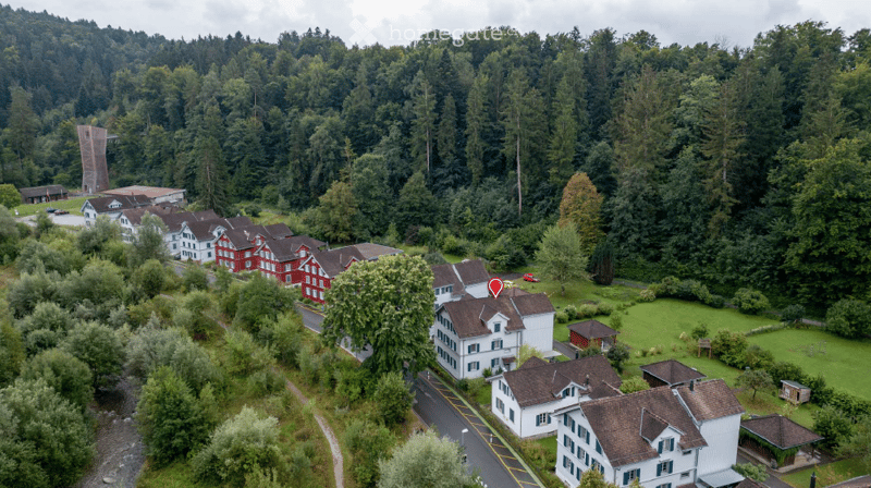Aerial view of a residential area with multiple houses, surrounded by lush green trees and a forested backdrop. The houses are mostly two-story structures with pitched roofs, and the area includes a small parking lot and a pathway.