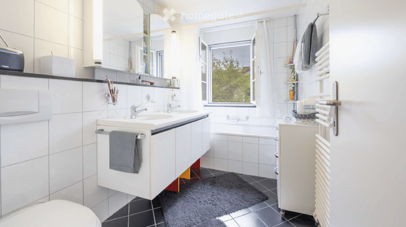 Modern bathroom with white tiles, double sink, mirror, bathtub, radiator, and towel rack. Features a large window with a view of greenery.