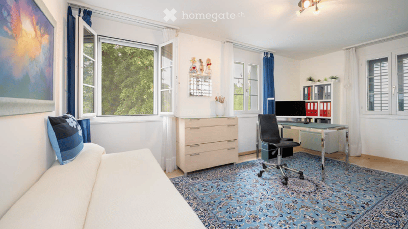 A well-lit room with a sofa, desk, chair, and cabinet. The room features large windows with blue curtains, a patterned rug, and a vase on the cabinet.