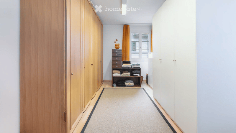 A narrow hallway with light wooden wardrobes on both sides, a small shelf unit with folded clothes and a vase, and a light-colored runner rug.