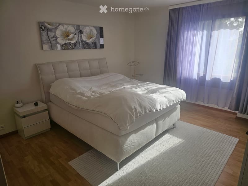 Well-lit bedroom with a large bed, nightstand, painting, and a lamp near the window.