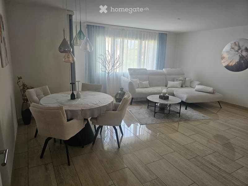 Modern living and dining area with a white sofa, coffee table, dining table, and chairs. The space features large windows with sheer curtains, pendant lights, and decorative vases.