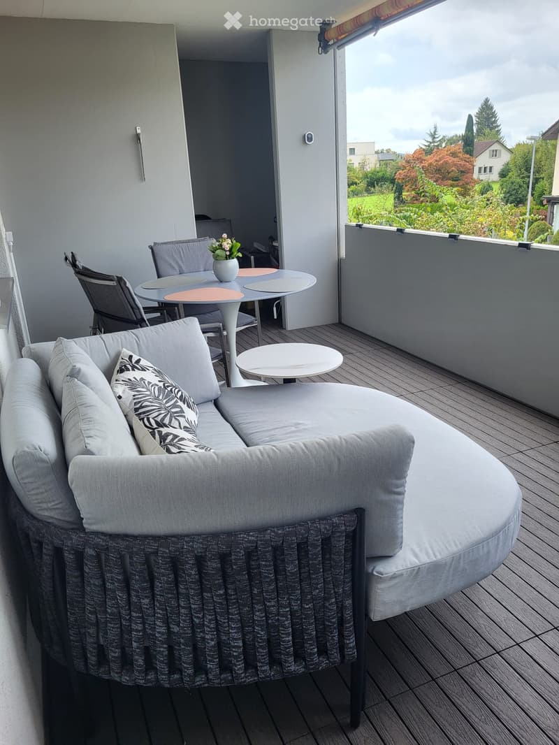 Balcony with wooden decking, grey sofa with cushions, small round table, chairs, and a flower pot.