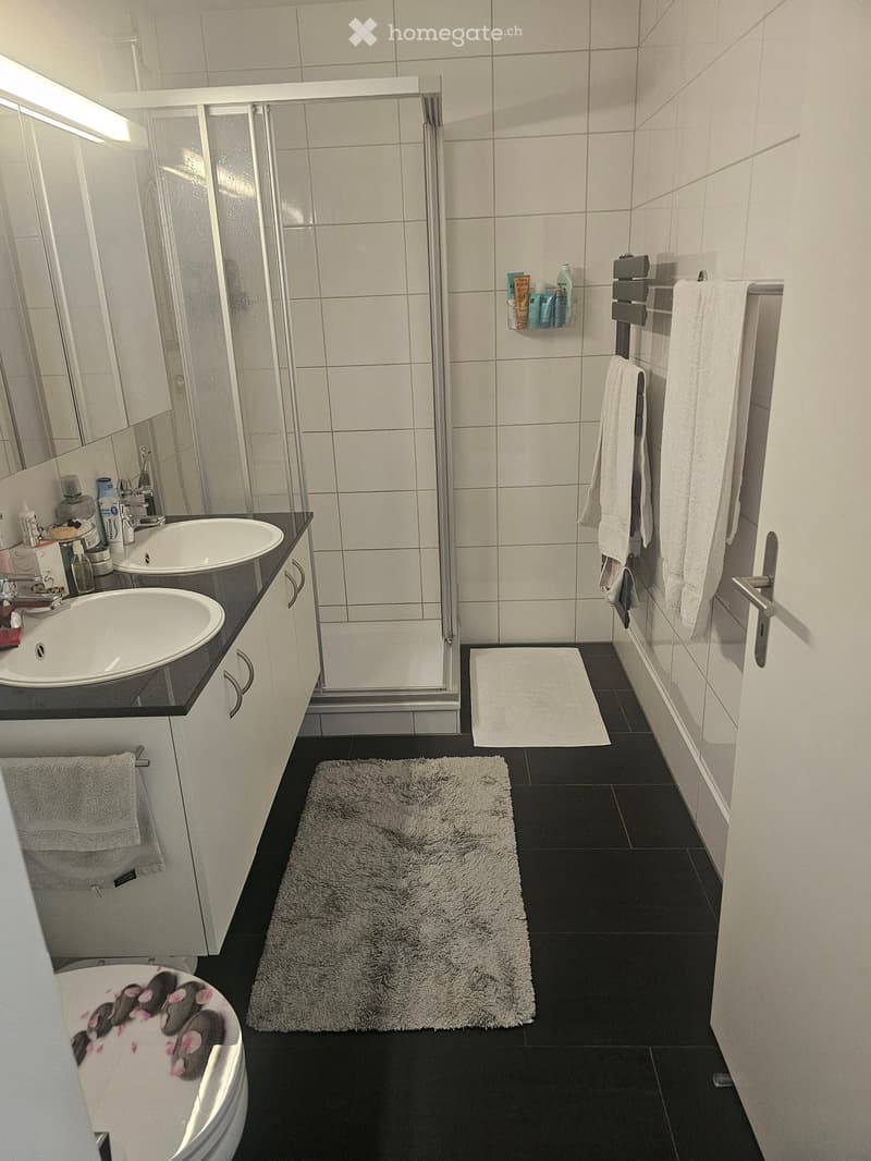 Modern bathroom with white tiles, black floor, double sink, shower, toilet, and towels.