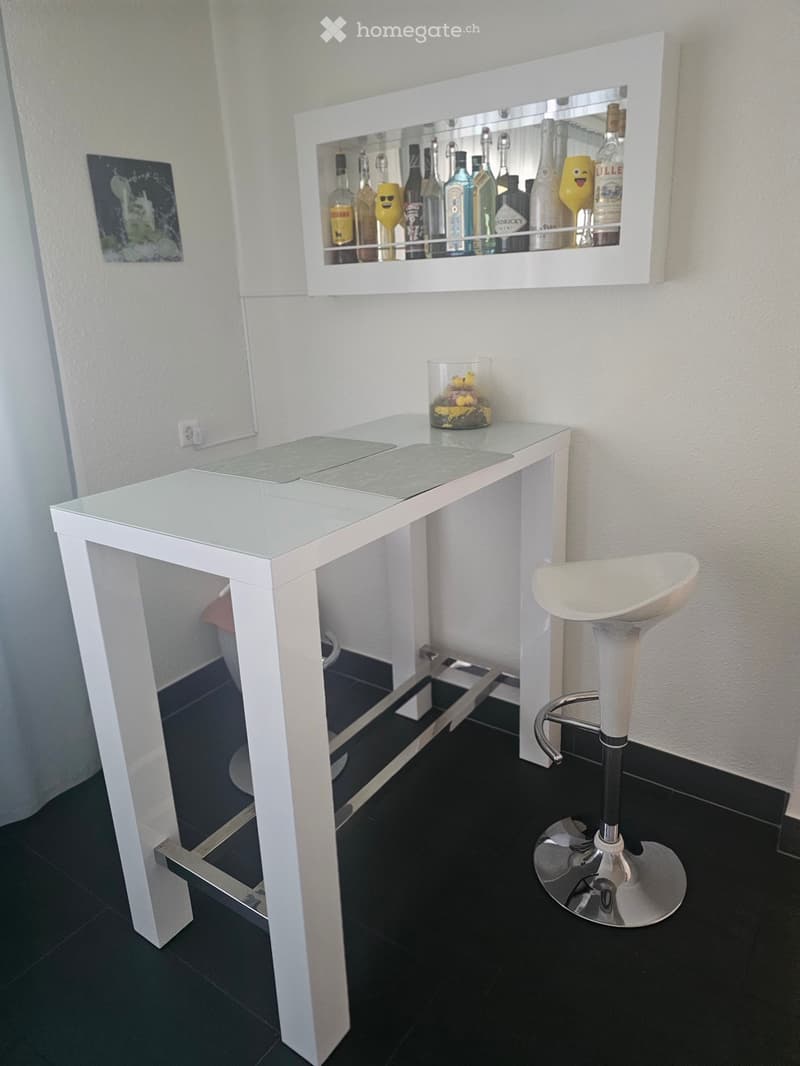 Modern white bar table with a mirrored backsplash displaying various bottles, accompanied by a white bar stool.