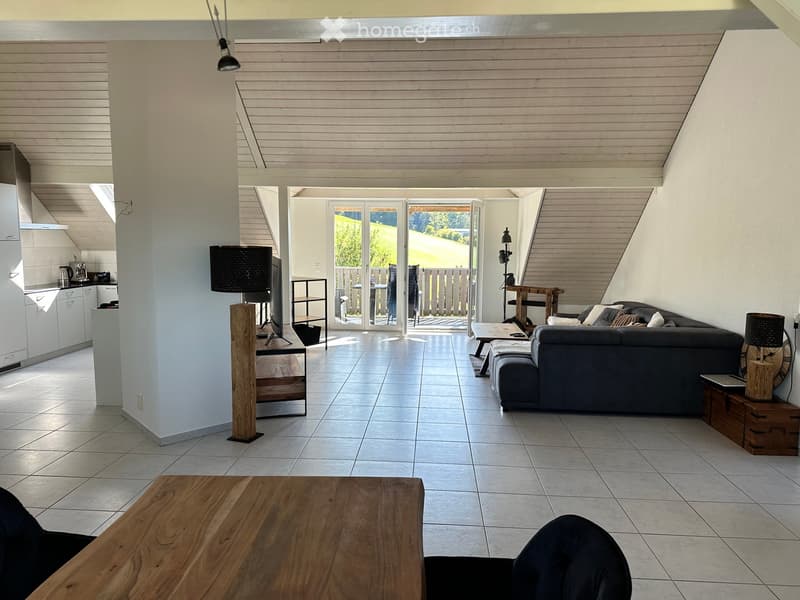 Open-plan living and dining area with a grey sofa, wooden dining table, TV, and kitchen area with white cabinets and appliances.