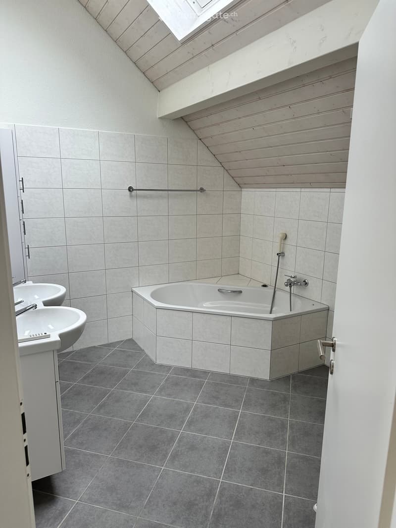 Modern bathroom with a large bathtub, two sinks, and grey tiled walls and floor. Features a skylight window and a wooden slanted ceiling.
