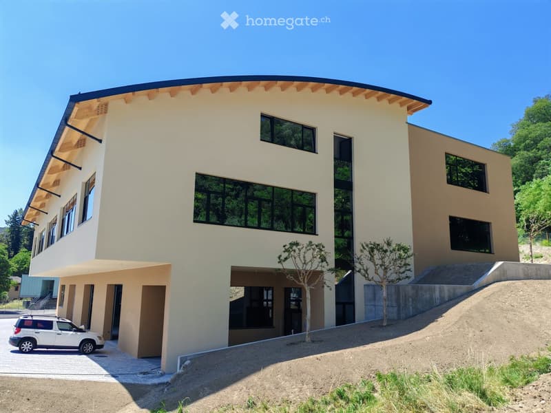 Modern two-story house with light beige walls, large windows, and a wooden roof overhang. Features a raised ground floor, a small parking area, and landscaped surroundings with trees and greenery.