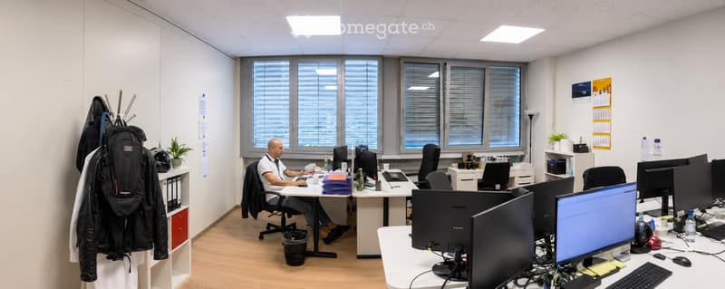 Office space with multiple desks, computer monitors, chairs, and a person working. Includes a backpack, plant, calendar, and filing cabinet.