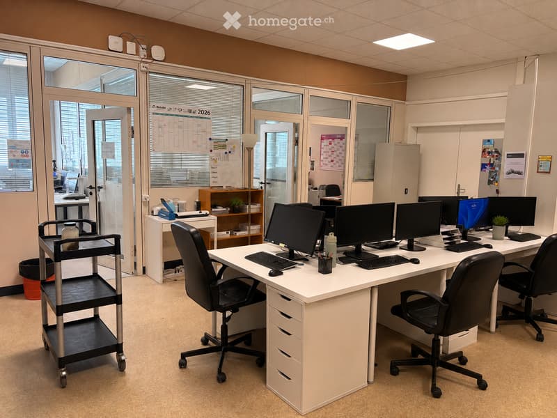 Office space with desks, chairs, multiple computer monitors, keyboards, and a small cart. Large windows with blinds and a calendar visible in the background.