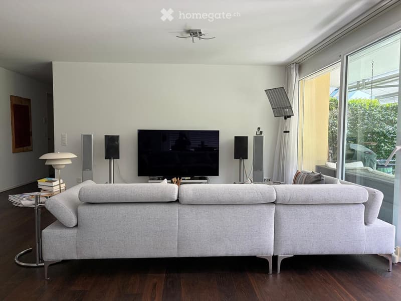 Modern living room with a grey sofa, a glass coffee table with books, a floor lamp, and a large television flanked by speakers. Large windows with white curtains let in natural light.