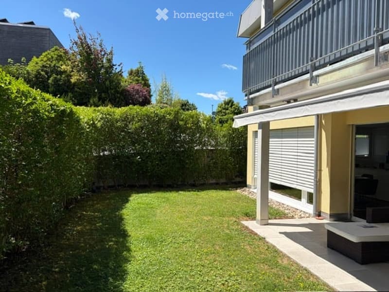 Well-maintained garden with neatly trimmed hedges, a small lawn, and a modern house with a balcony and covered patio area.