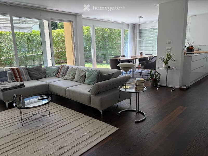 Modern living room with a grey sectional sofa, coffee tables, floor lamp, dining area with a table and chairs, and a kitchen counter with a potted plant.