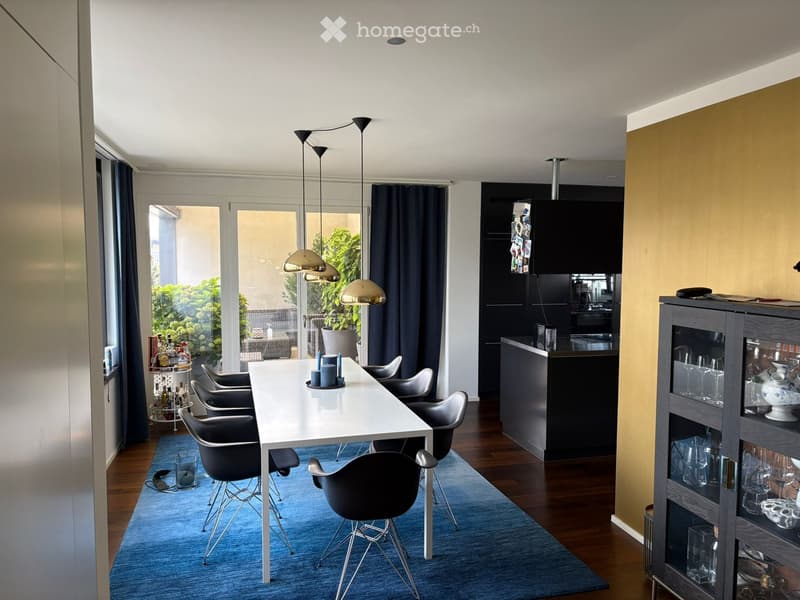 Modern dining area with a white dining table, six black chairs, pendant lights, a blue rug, and a bar cart with bottles. A cabinet and shelf are visible, along with a large window with dark curtains.