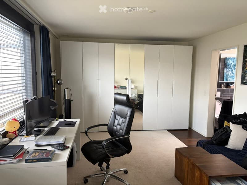 A well-furnished room with a desk, chair, sofa, and a large window with blinds. The room features a modern aesthetic with a mix of wooden and white furniture.