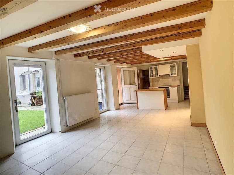 Bright, open-plan living and dining area with exposed wooden beams on the ceiling, large sliding glass doors leading to a garden, and a modern kitchen area with white cabinets and a central island.