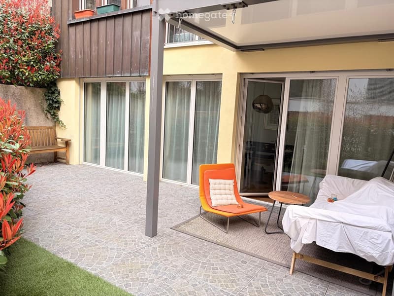 Balcony with tiled floor, orange chair, small table, covered sofa, wooden bench, and potted plants.