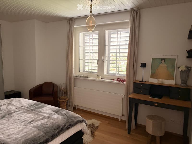 Cozy bedroom with a large bed, a brown armchair, a wooden desk with a lamp, a stool, and a picture frame on the wall. The room has large windows with curtains and a hanging light fixture.