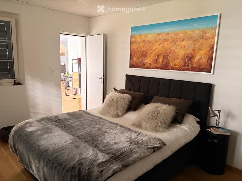 Modern bedroom with a large bed, fur throw, decorative pillows, nightstand, and a large abstract painting on the wall. An open doorway leads to a kitchen area with a table and chairs.