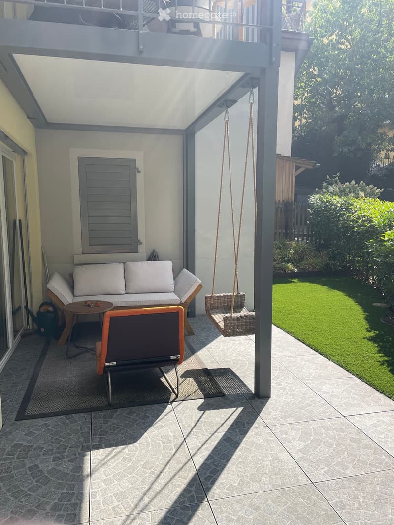 Balcony with a sofa, small table, chair, and hanging swing, overlooking a well-maintained garden.